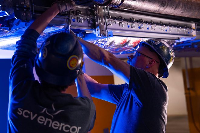 Deux techniciens de SCV Enercor en casque de sécurité installent un système de rail métallique au plafond dans un environnement industriel éclairé en bleu