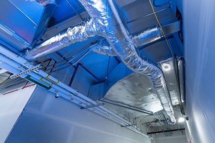 Conduits de ventilation gainés en aluminium installés au plafond d’un corridor technique, avec éclairage bleuté et murs en plaques de plâtre