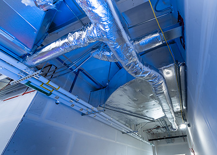 Conduits de ventilation gainés en aluminium installés au plafond d’un corridor technique, avec éclairage bleuté et murs en plaques de plâtre