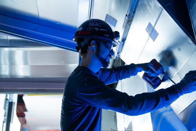Technicien en ventilation installant un conduit métallique dans un bâtiment commercial, portant un casque de sécurité et utilisant une visseuse, éclairage industriel bleu