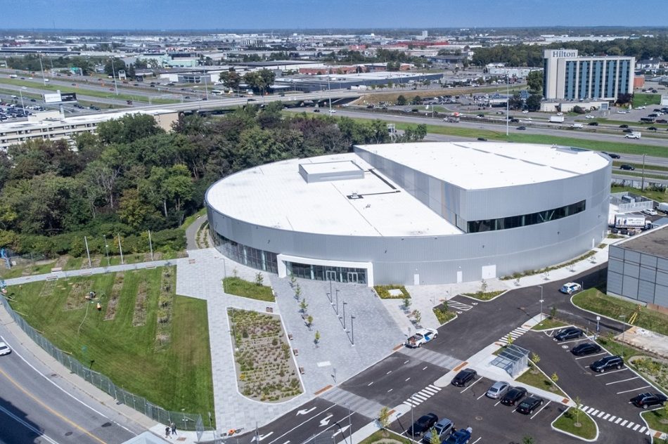 Vue aérienne du Complexe aquatique de Laval, montrant le bâtiment moderne à toit blanc, les aménagements paysagers, le stationnement et l’autoroute en arrière-plan