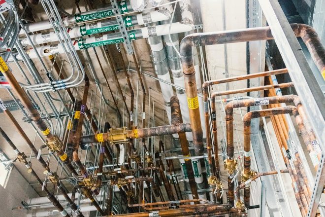 Dense network of copper piping and mechanical ducts suspended from the ceiling of a technical room, with labels for medical air and hot water
