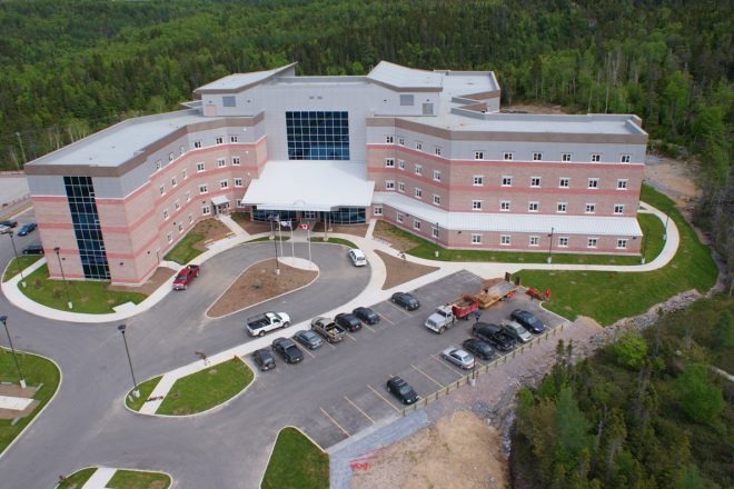 Aerial view of the completed Corner Brook hospital, modern brick and metal panel building surrounded by forest, with visible parking and main entrance