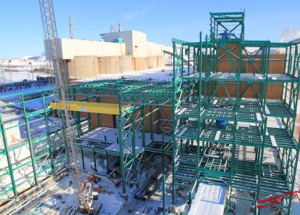 Structure d'acier verte en construction sur le site minier de Mont-Wright en hiver, avec grue, neige au sol et bâtiments industriels en arrière-plan