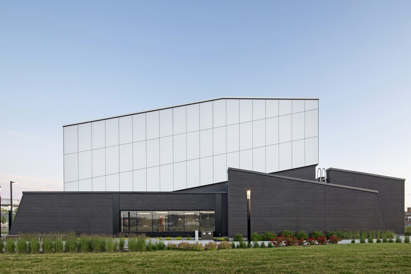 Exterior view of the STM Côte-Vertu station, featuring a modern building with a textured black façade, a geometric white upper volume, and well-maintained landscaping