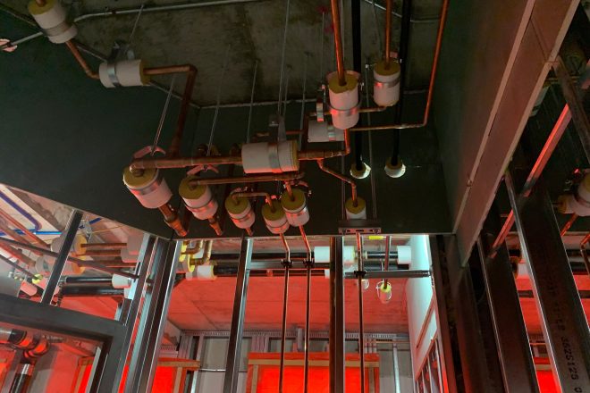 Complex network of copper and PVC pipes with insulation, suspended from the ceiling of a building under construction, lit by temporary red lighting