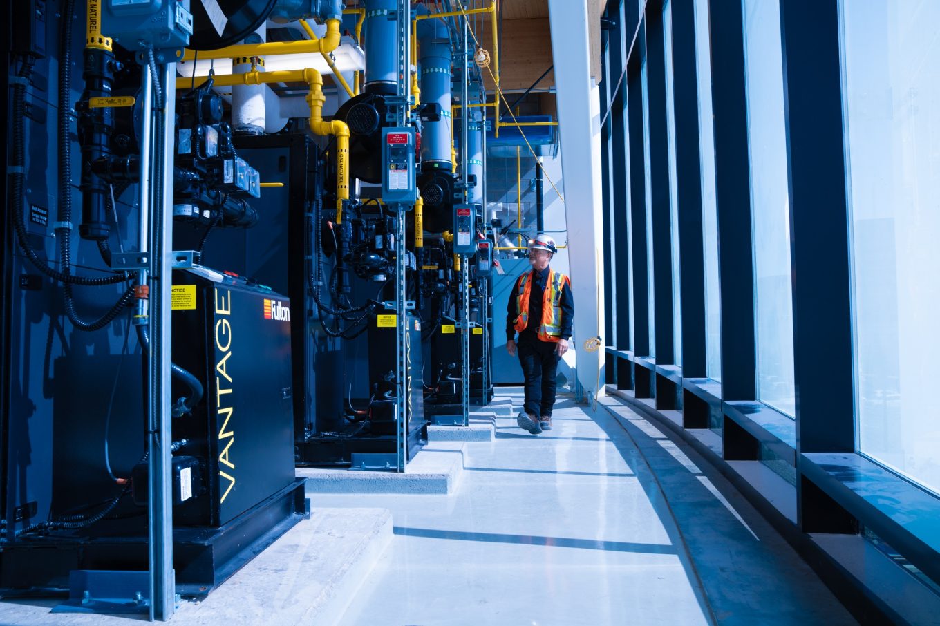A worker in a safety vest inspects a row of Fulton VANTAGE industrial mechanical units in a glass-enclosed technical room