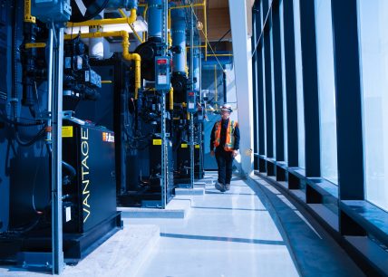 Un travailleur en gilet de sécurité inspecte une rangée d’équipements mécaniques industriels Fulton VANTAGE dans une salle technique vitrée