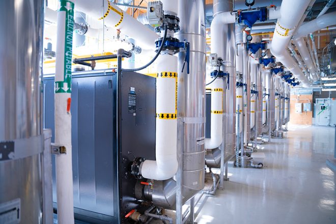 Salle mécanique lumineuse avec une rangée de conduites isolées pour l’eau réfrigérée et l’eau chaude, vannes motorisées et équipements industriels en acier inoxydable, installés sur un plancher lustré