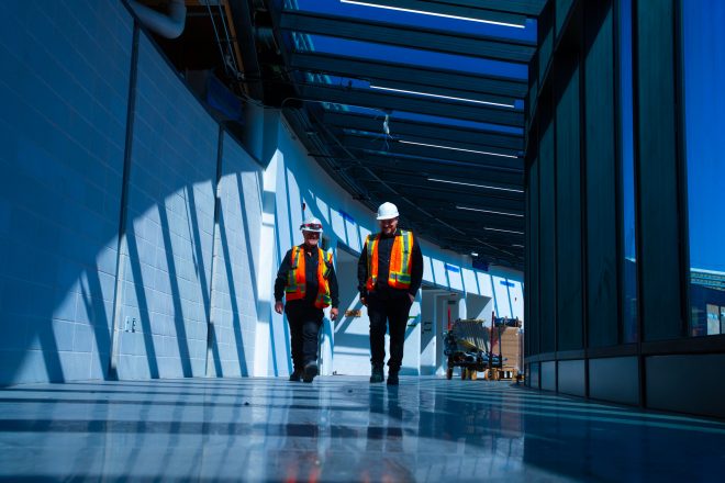 Two workers wearing hard hats and safety vests walk through a glass corridor of a building under construction, illuminated by natural light