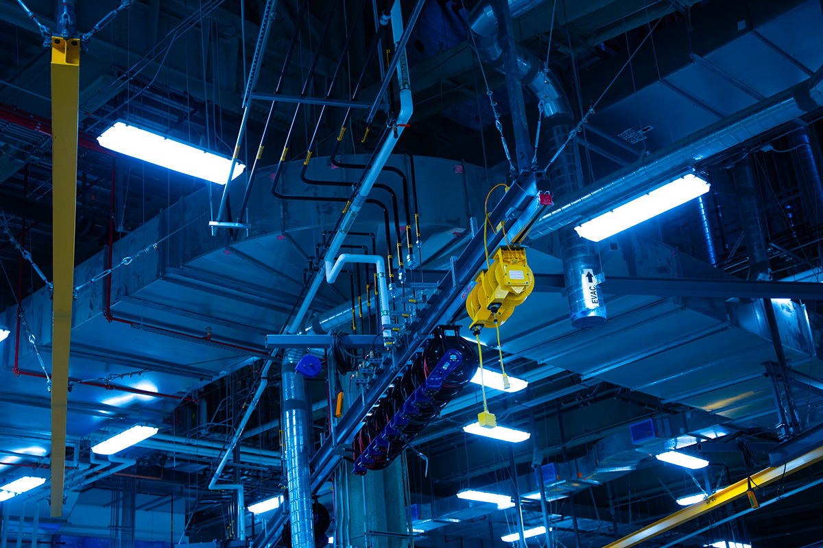 Plafond d’une salle mécanique industrielle avec tuyauterie étiquetée, enrouleurs de câbles suspendus, systèmes de ventilation et éclairage fluorescent bleuté