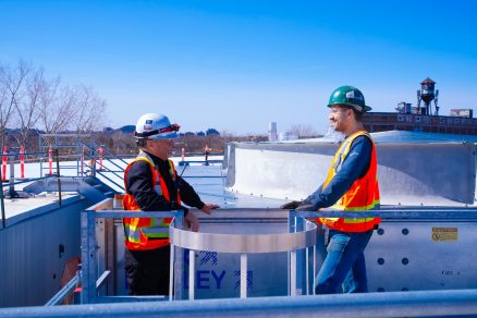 Two construction workers having a discussion on an industrial rooftop while wearing full safety gear, including helmets, gloves, and reflective vests. The scene emphasizes teamwork, safety standards, and field coordination in the construction industry in Québec.