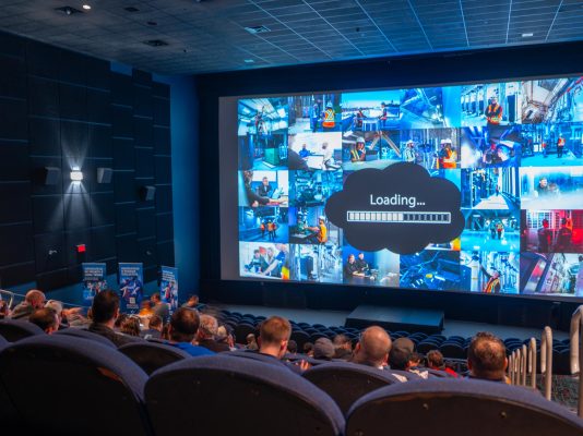 A packed movie theater hosting a corporate presentation with a large screen showing a loading bar and a collage of employees on job sites. The event showcases employee recognition, team spirit, and corporate culture in the industrial and engineering sector.